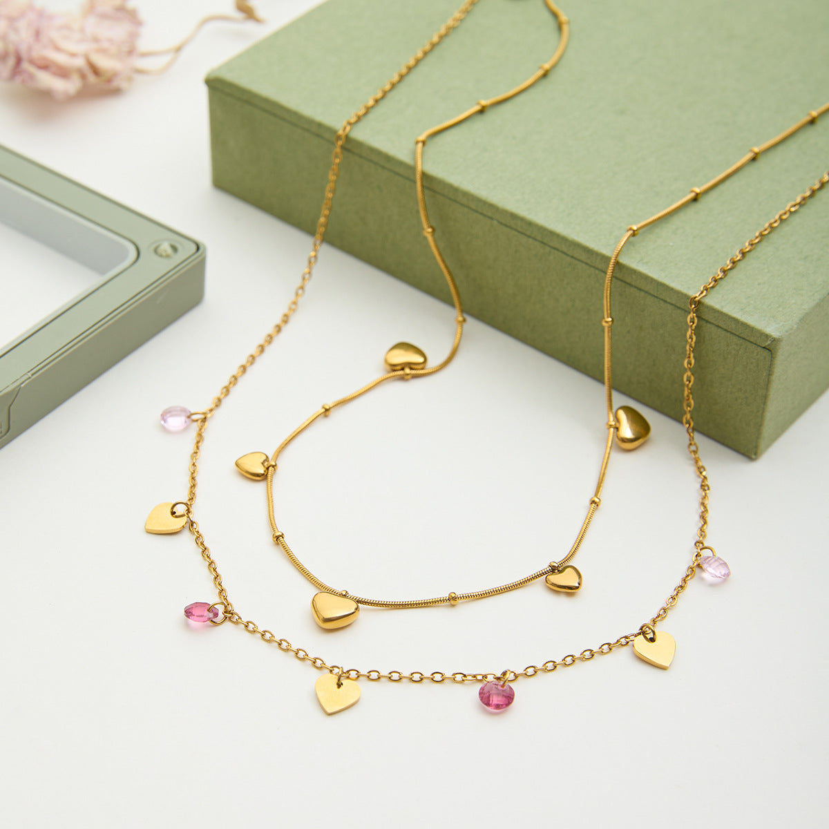 Gold necklace with heart and pink bead charms on a white surface with a green box in the background.