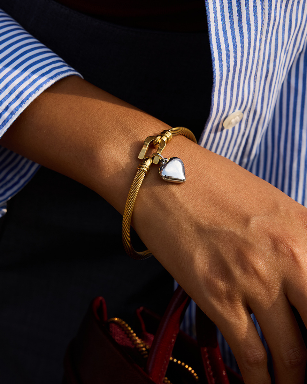 Gold bracelet with a heart charm on a wrist, wearing a blue and white striped shirt.