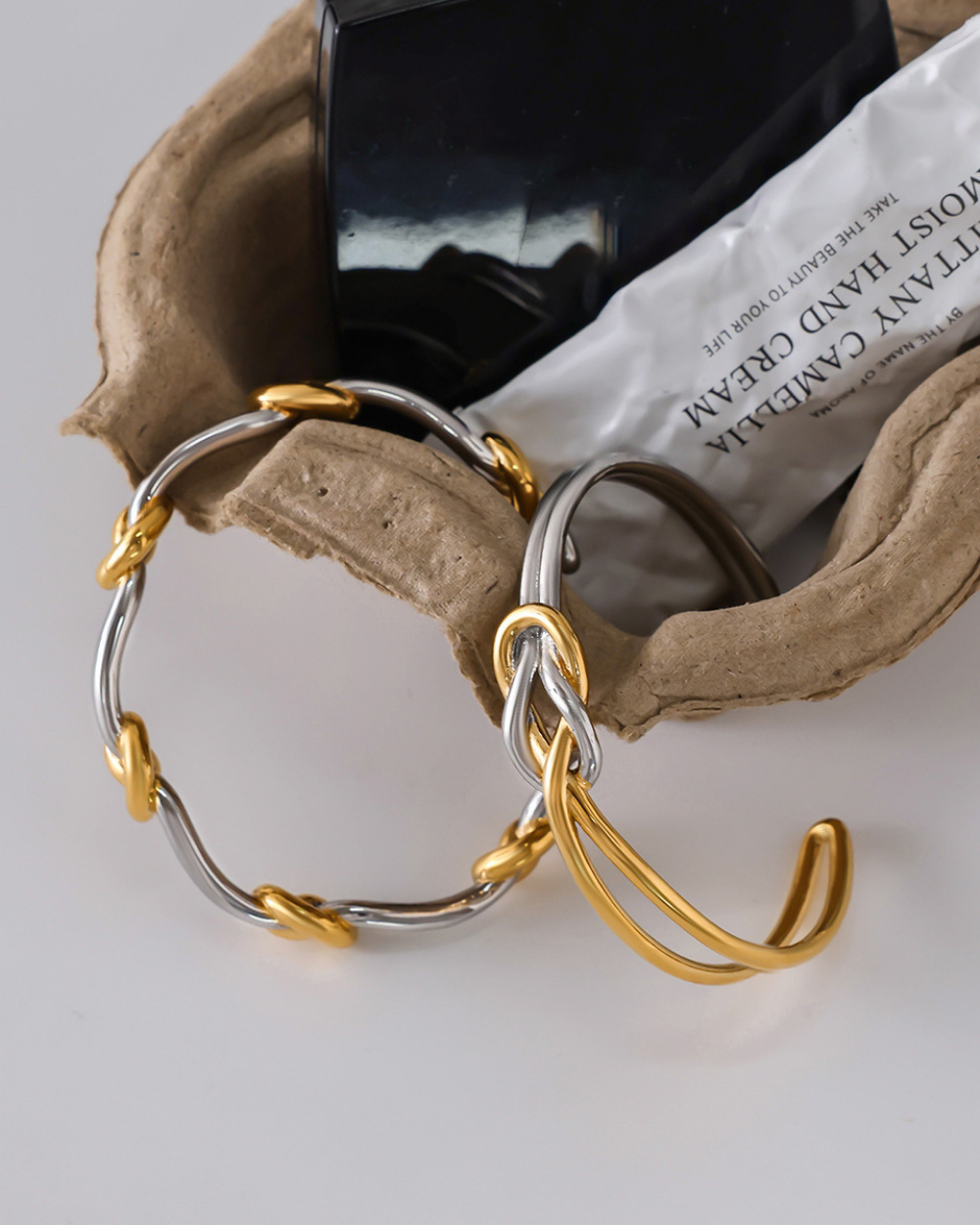 Gold and silver hoop earrings on a textured surface with a black container and white paper in the background.