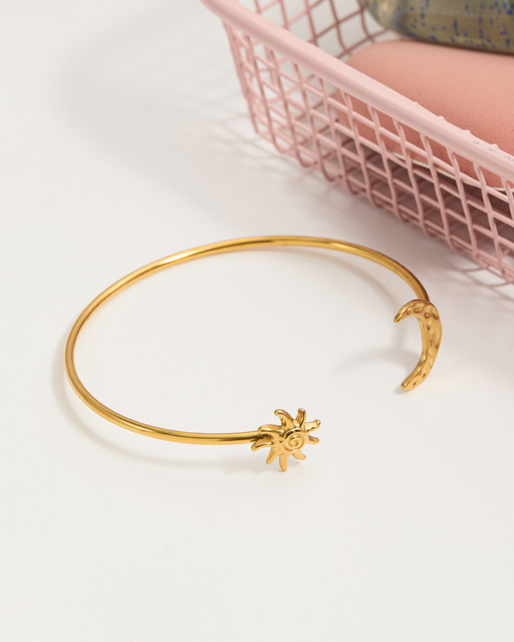 Gold bracelet with a sunflower charm on a white surface with a pink basket in the background.