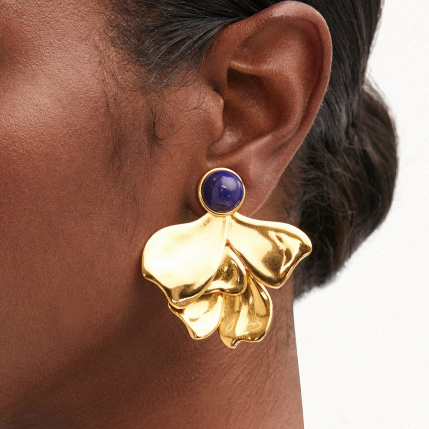 Model wearing bold gold petal earrings with blue stone detail