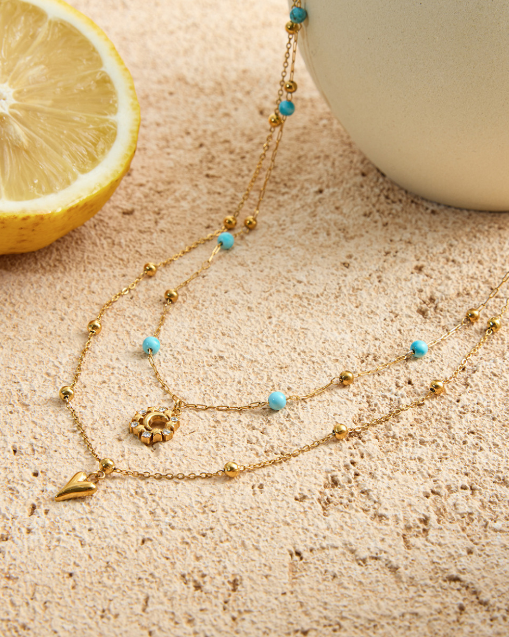 Gold necklace with blue beads on a textured surface with a lemon and white object in the background