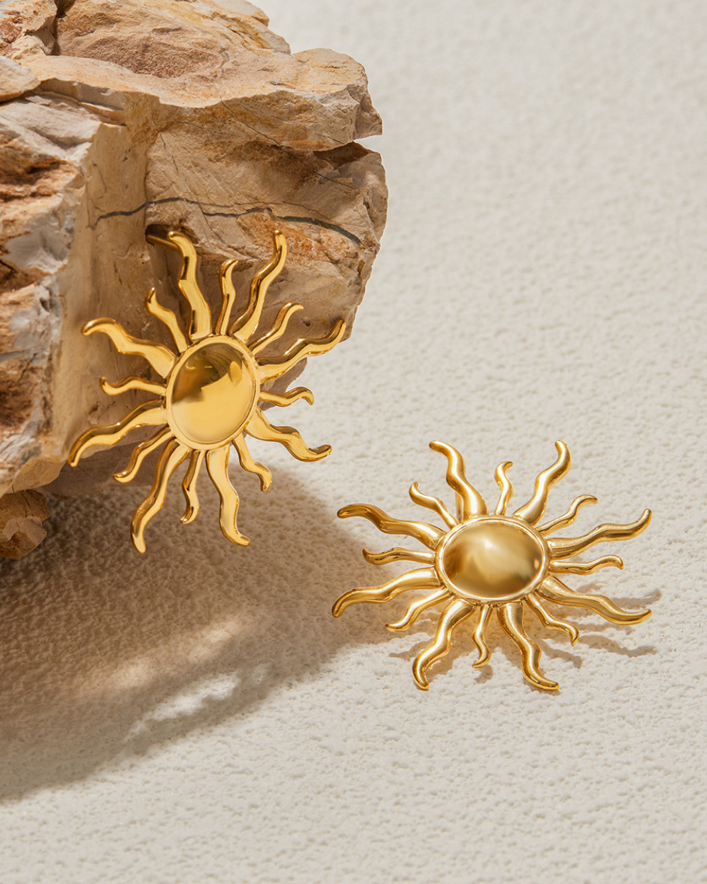 Gold sun-shaped earrings on a textured beige surface with a stone background