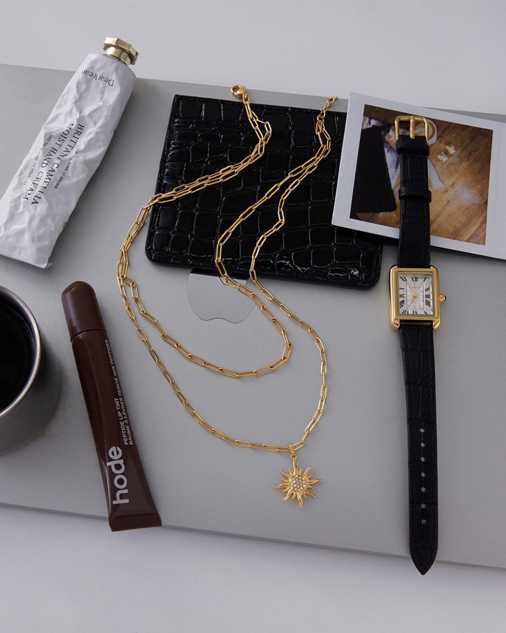 Gold necklace with a sun pendant, black watch, and coffee on a gray surface.
