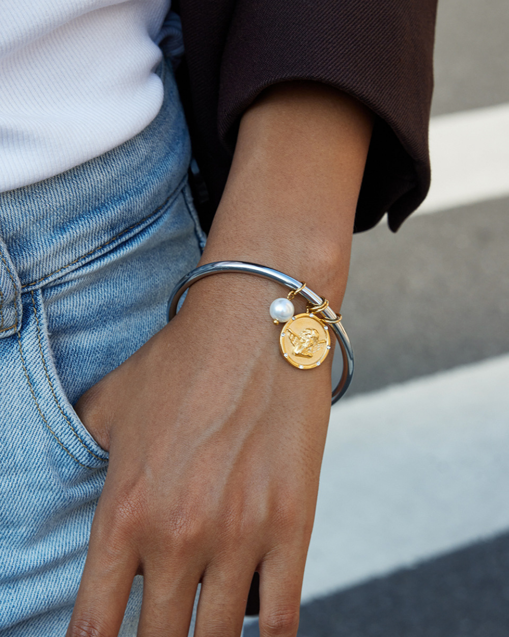 Close-up of a hand wearing a silver bracelet with a gold coin charm and pearl on a blurred background.