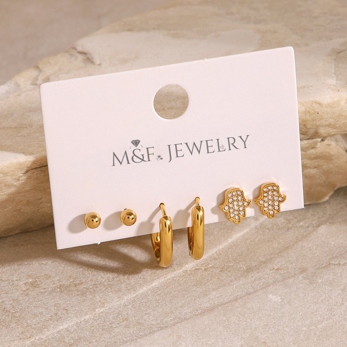 Gold-plated Hamsa hand earrings with clear crystals, laid flat on beige stone for lifestyle appeal.