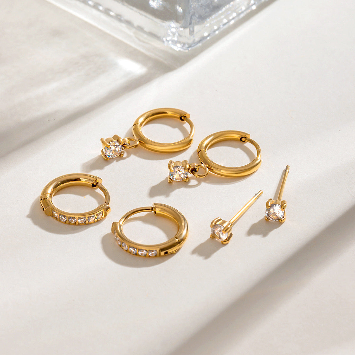 Close-up of gold huggie earrings with dangling round zircon charm on soft fabric background.