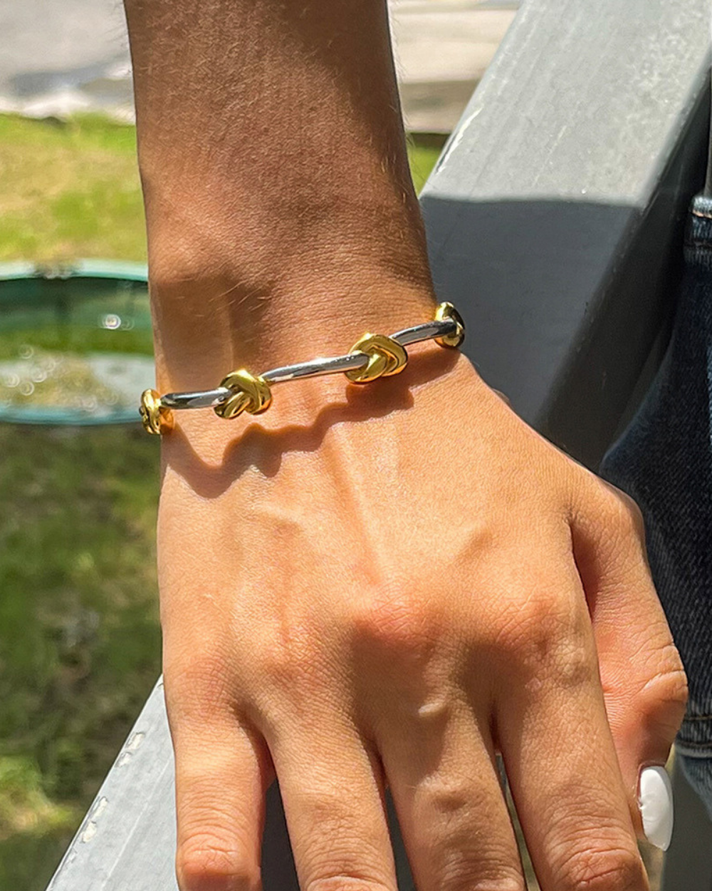 Hand wearing a gold and silver bracelet with a blurred outdoor background
