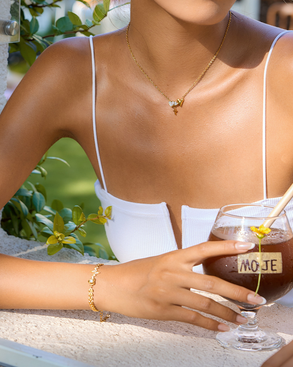 Woman holding a mojito cocktail with a gold necklace and bracelet, outdoor setting