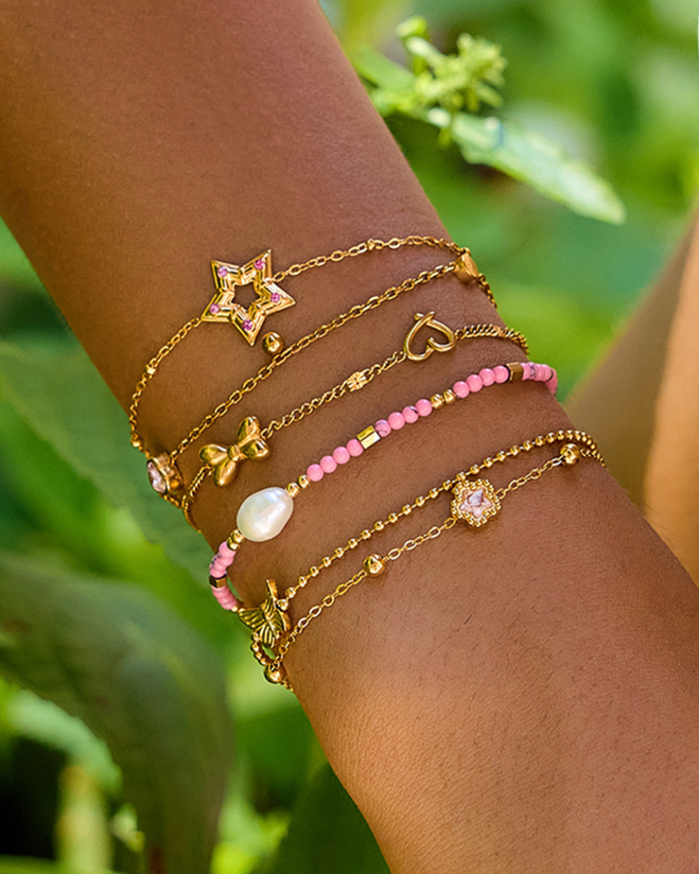 Multiple gold and pink bracelets on a wrist with a green leafy background