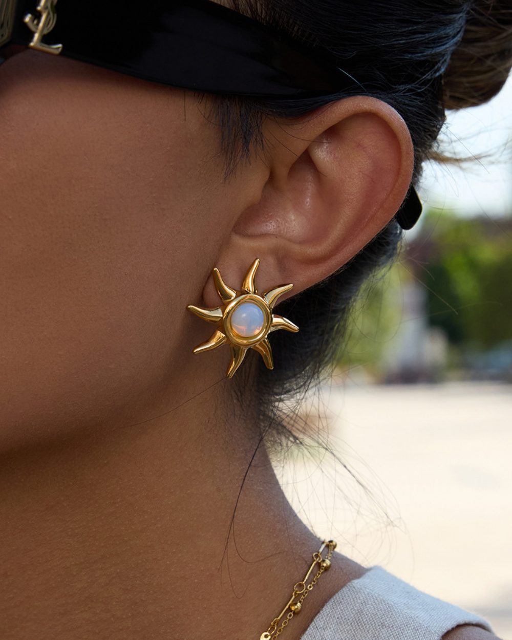 Close-up of a person wearing a gold sun-shaped earring with a blurred outdoor background