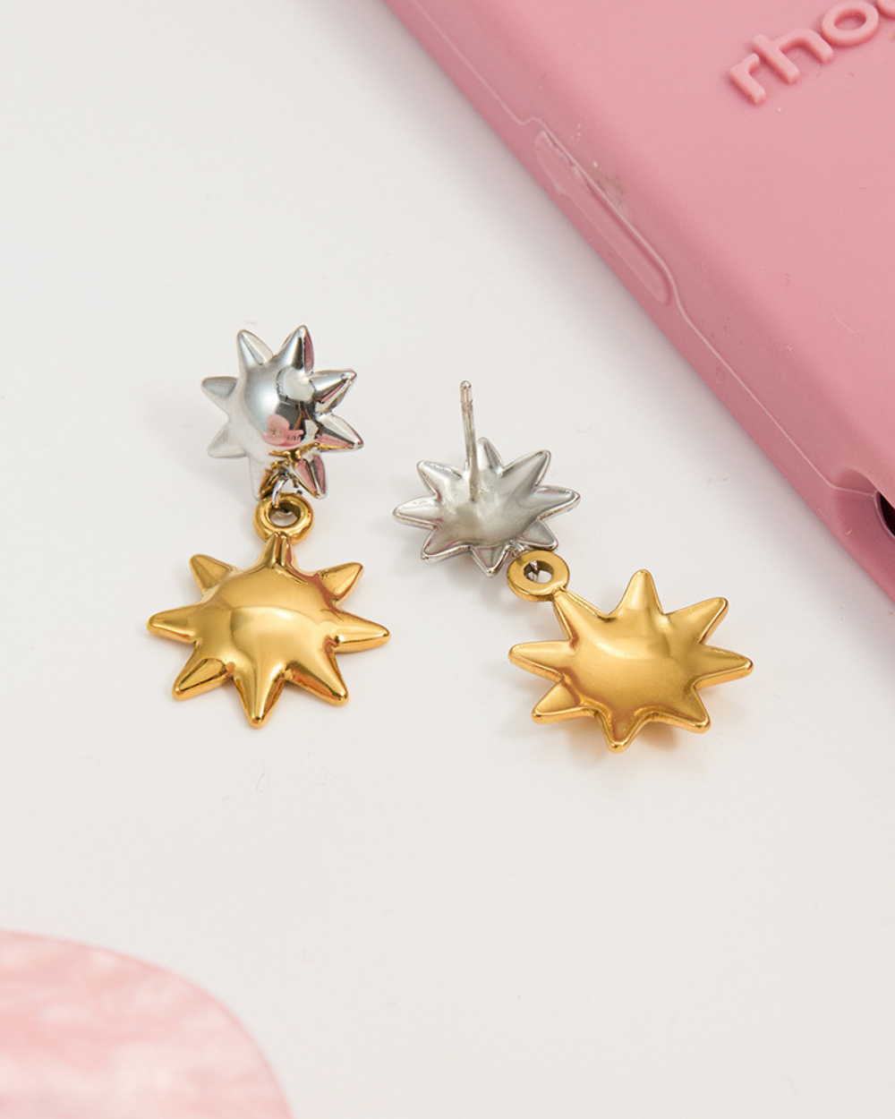 Pair of star-shaped earrings on a light pink background with a pink phone case.