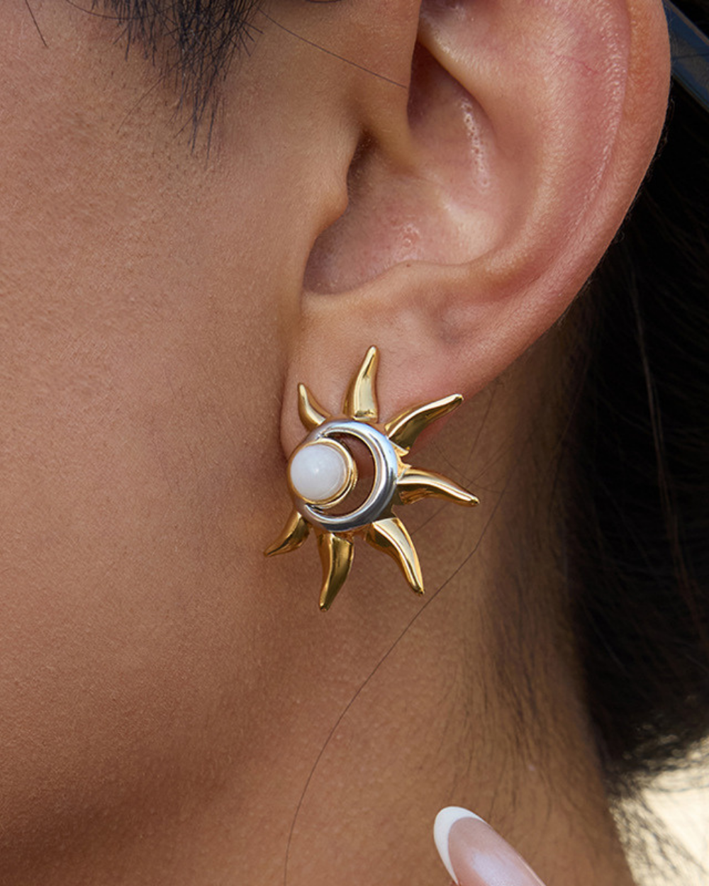Gold sun-shaped earring with a pearl center worn by a person.