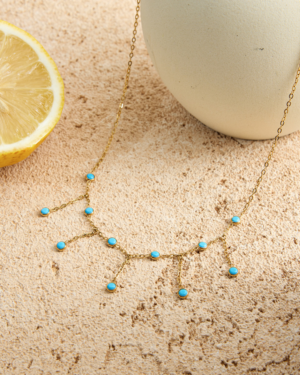 Gold necklace with turquoise stones on a textured surface next to a lemon and a white ball.