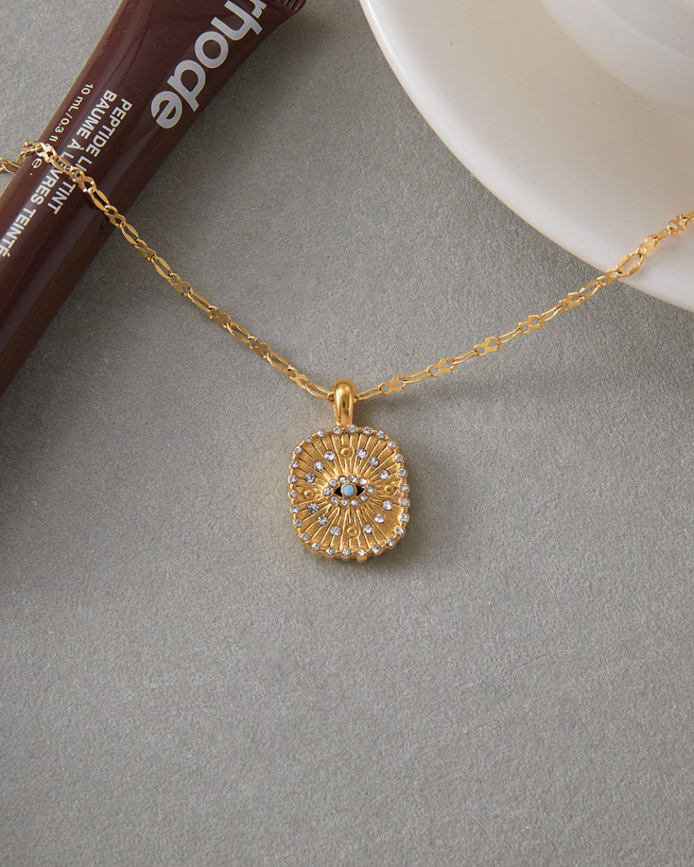 Lifestyle flatlay of gold sun charm necklace on grey surface next to coffee, modern jewelry aesthetic.