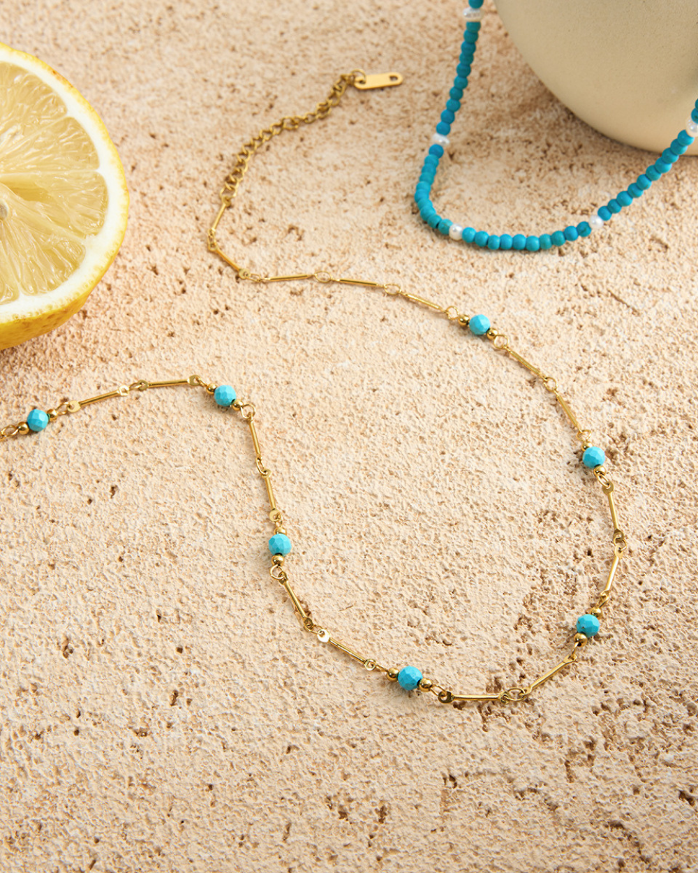Gold necklace with turquoise beads on a sandy surface next to a lemon slice.