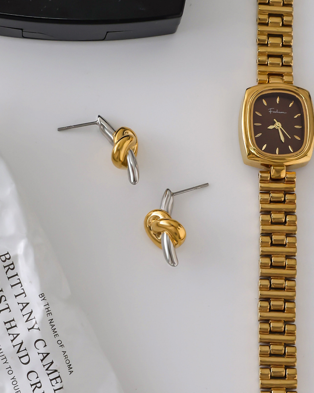 Gold and silver knot earrings with a gold watch on a white surface.
