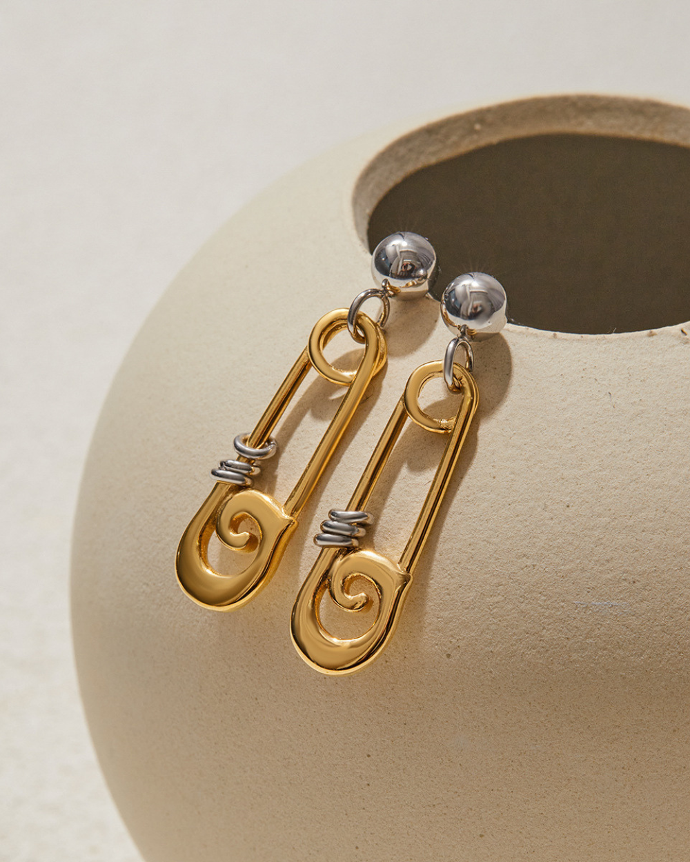 Gold and silver safety pin earrings on a beige stone surface