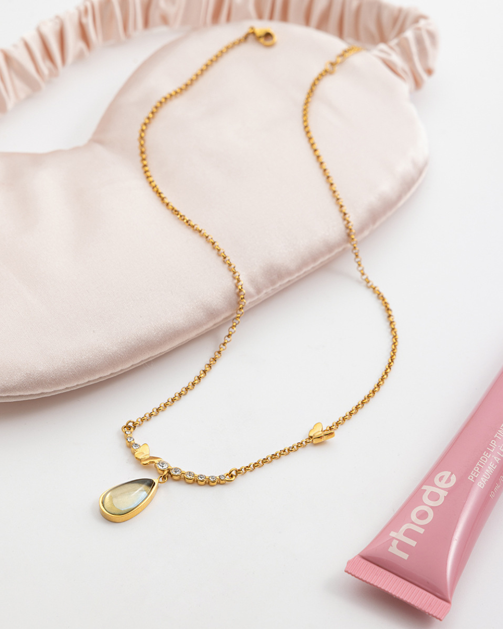 Gold necklace with a teardrop pendant on a pink cushion, with a pink tube of lip balm in the background.