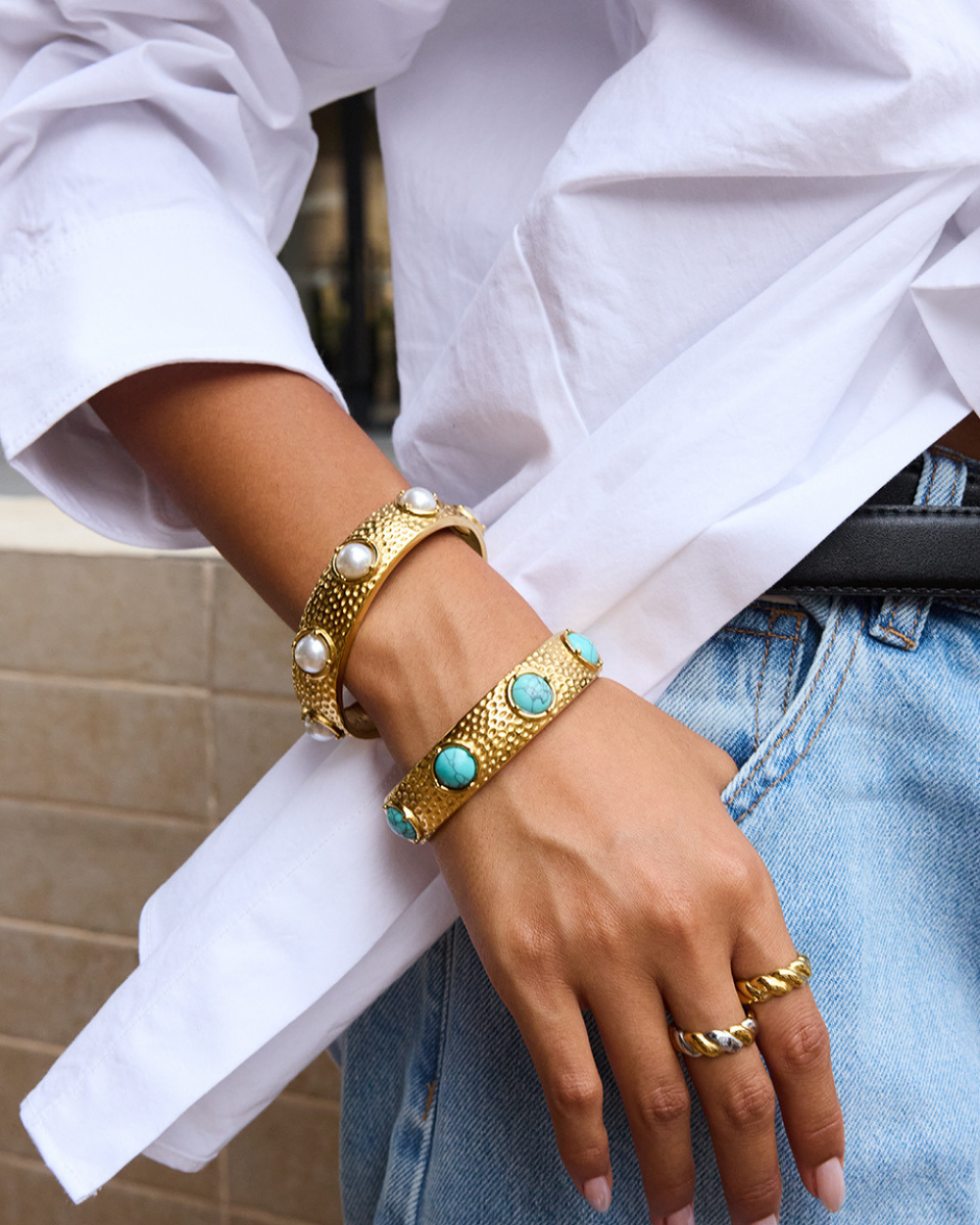 Close-up of a hand wearing gold bracelets and rings with a white shirt and denim jeans.