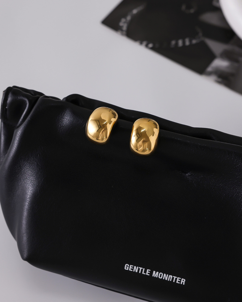 Black handbag with gold accents and 'GENTLE MONSTER' branding on a light gray background