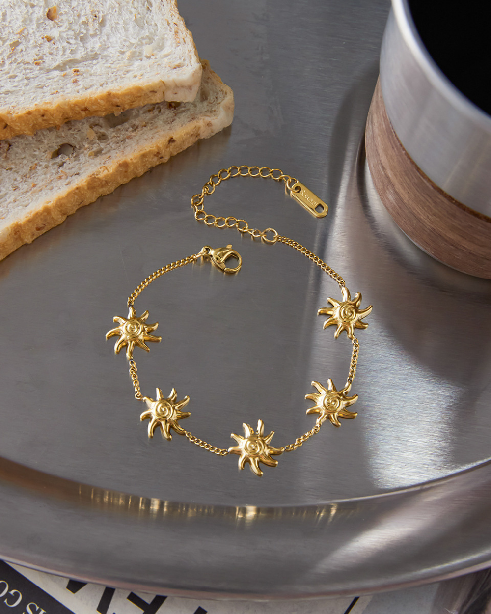 Gold bracelet with sun designs on a metallic surface next to toast and a cup.