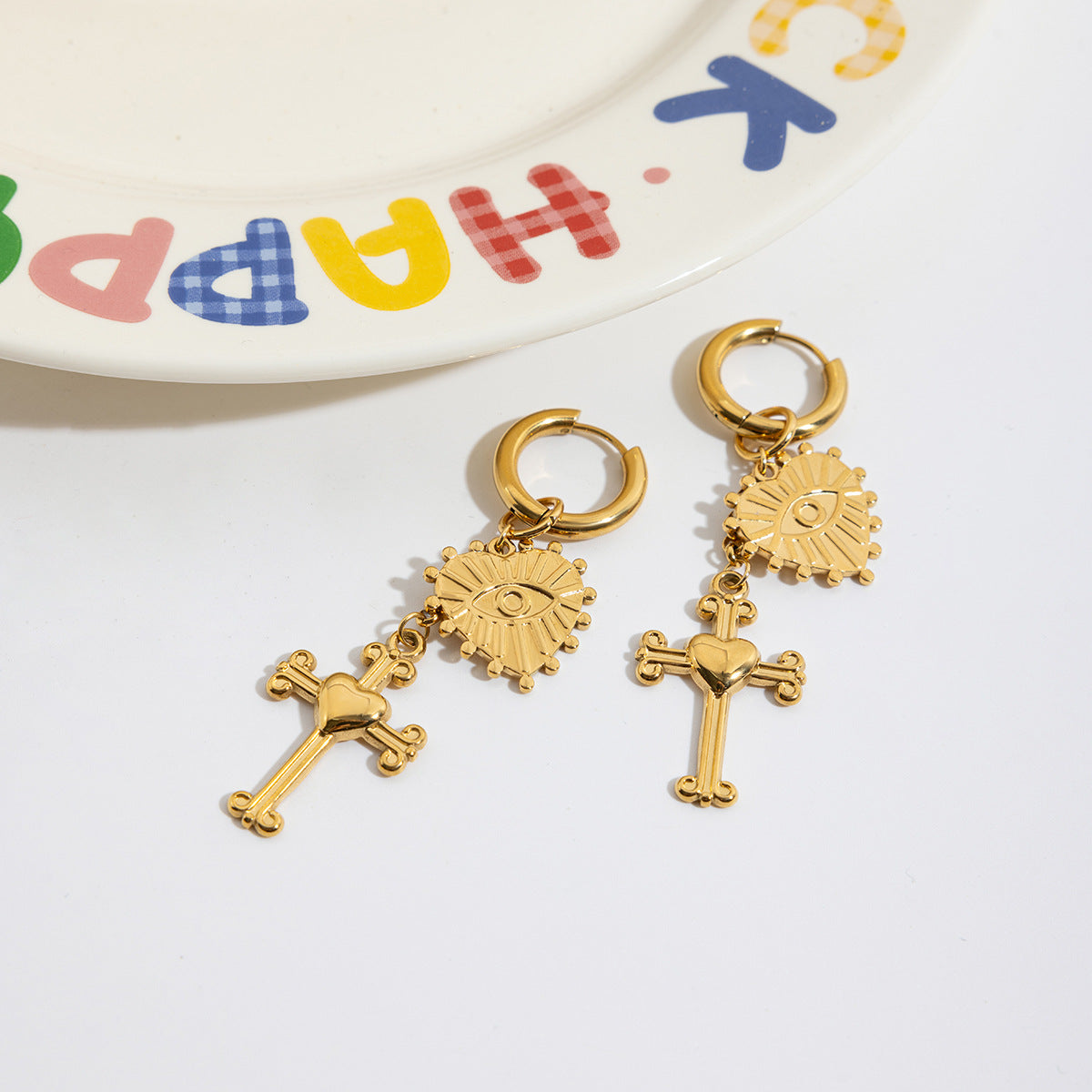 Gold earrings with religious charms on a white surface with a colorful 'Happy Birthday' plate in the background.