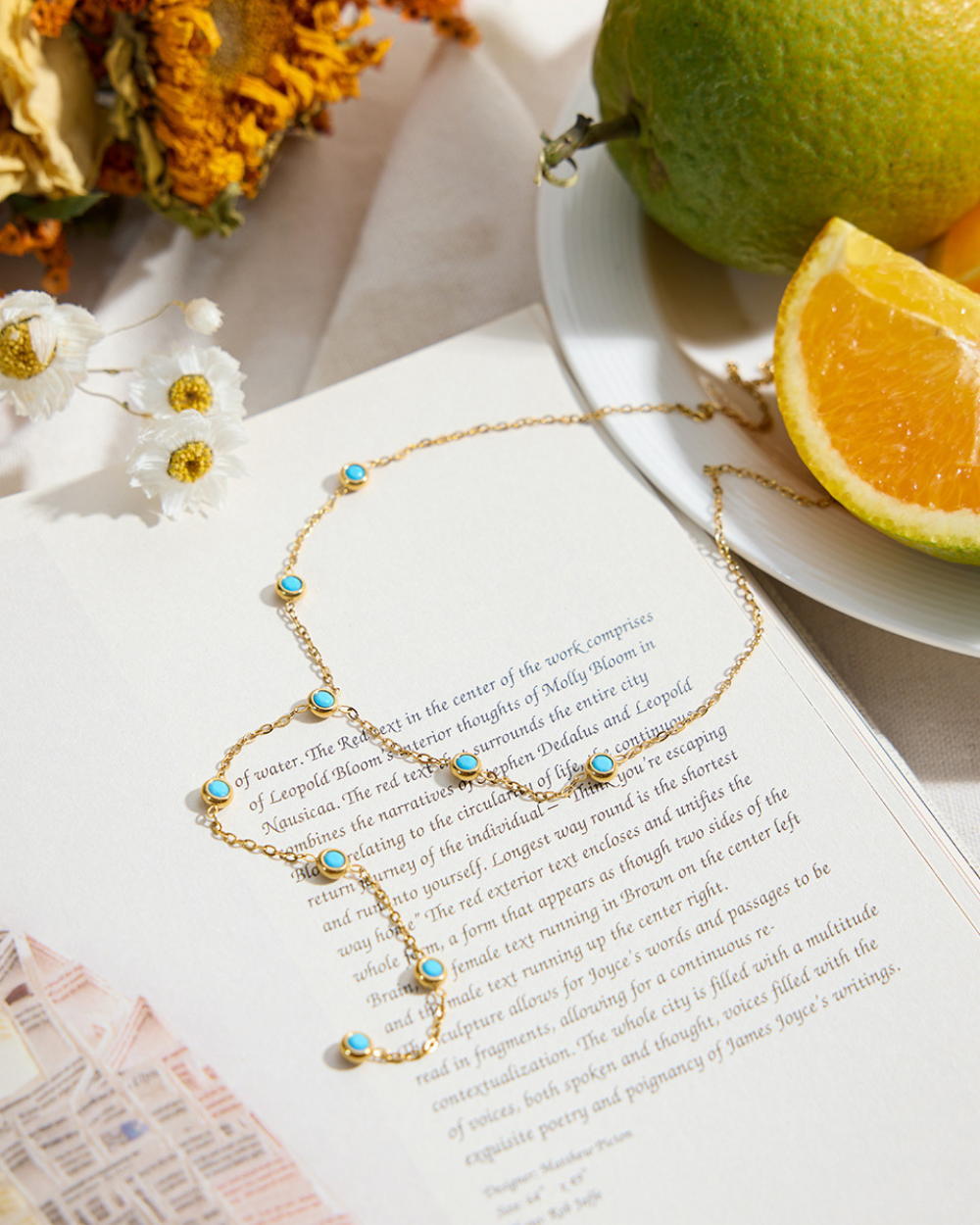 Gold necklace with turquoise stones on an open book with fruits and flowers in the background