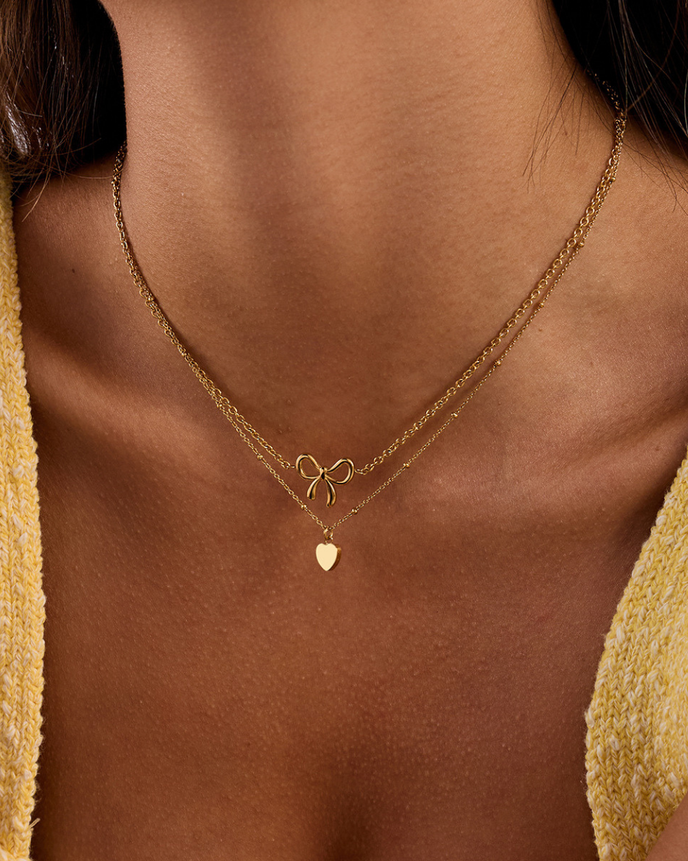 Close-up of a person wearing a gold necklace with a bow and heart pendant.