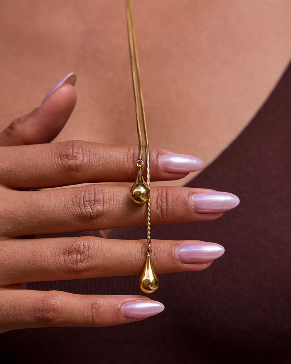 Gold teardrop-shaped pendant on a chain held by a hand with pink nail polish against a brown background.