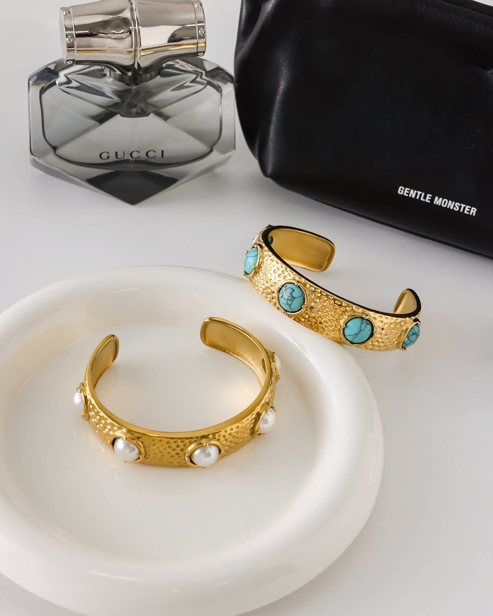 Gold bracelets with turquoise stones on a white surface, next to a Gucci perfume bottle and black handbag.
