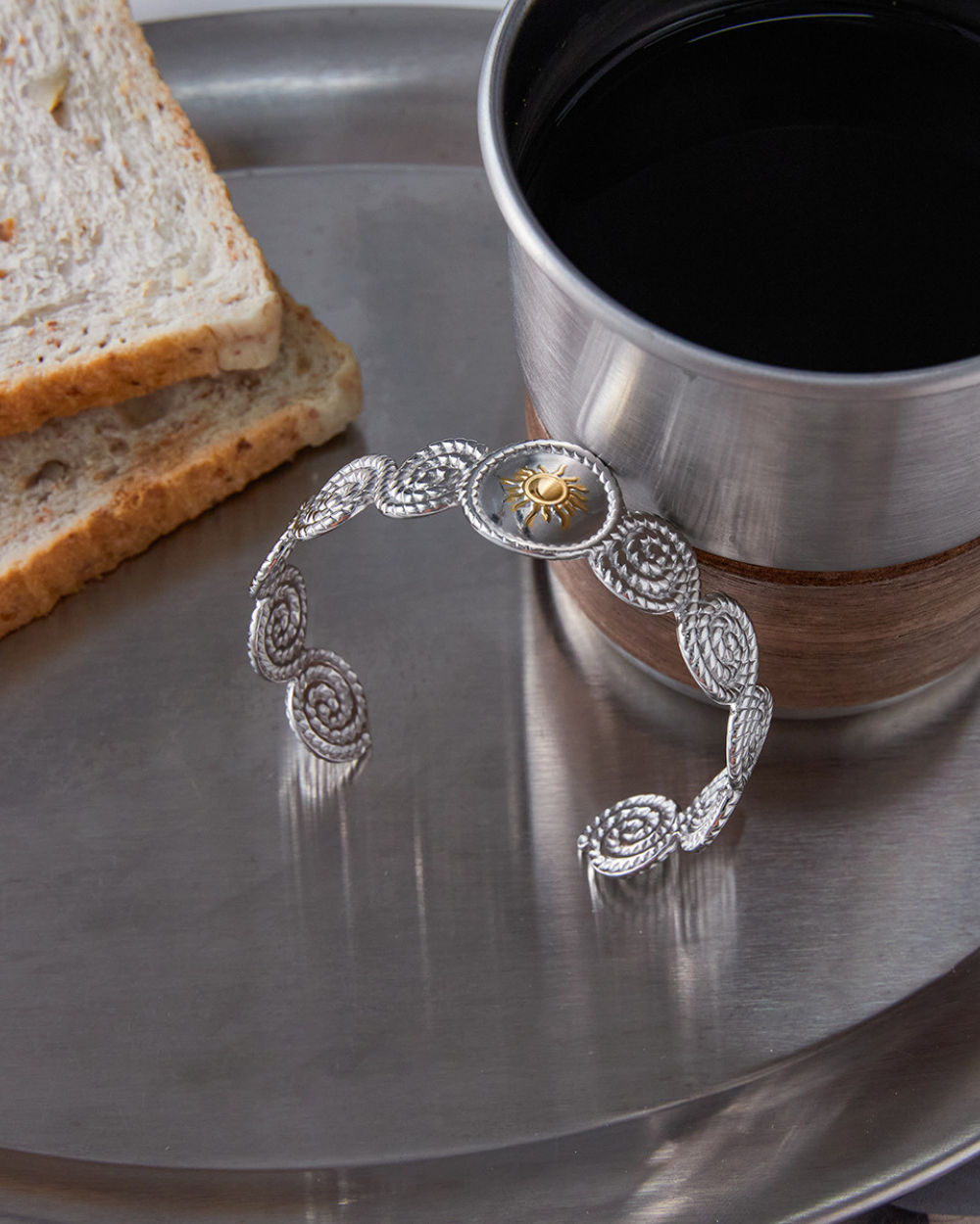 Silver bracelet on a metal surface with a cup of coffee and toast in the background