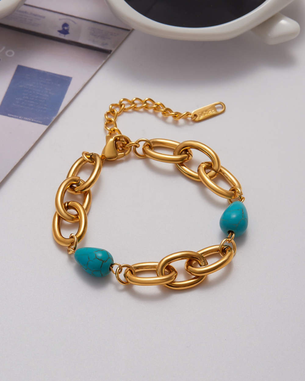 Gold chain bracelet with turquoise beads on a light gray surface.