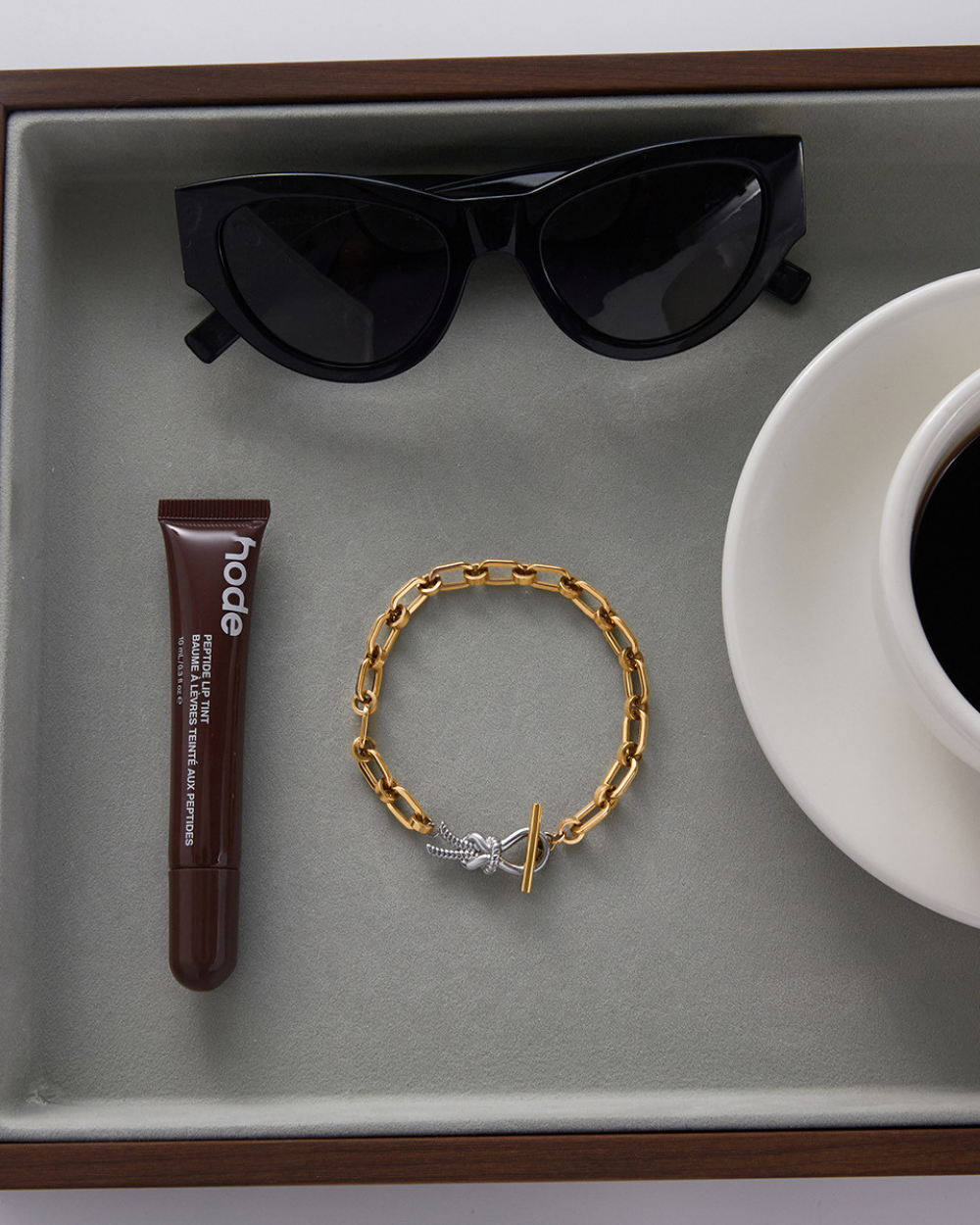 Gold bracelet, tube of lip balm, sunglasses, and cup of coffee on a gray tray.