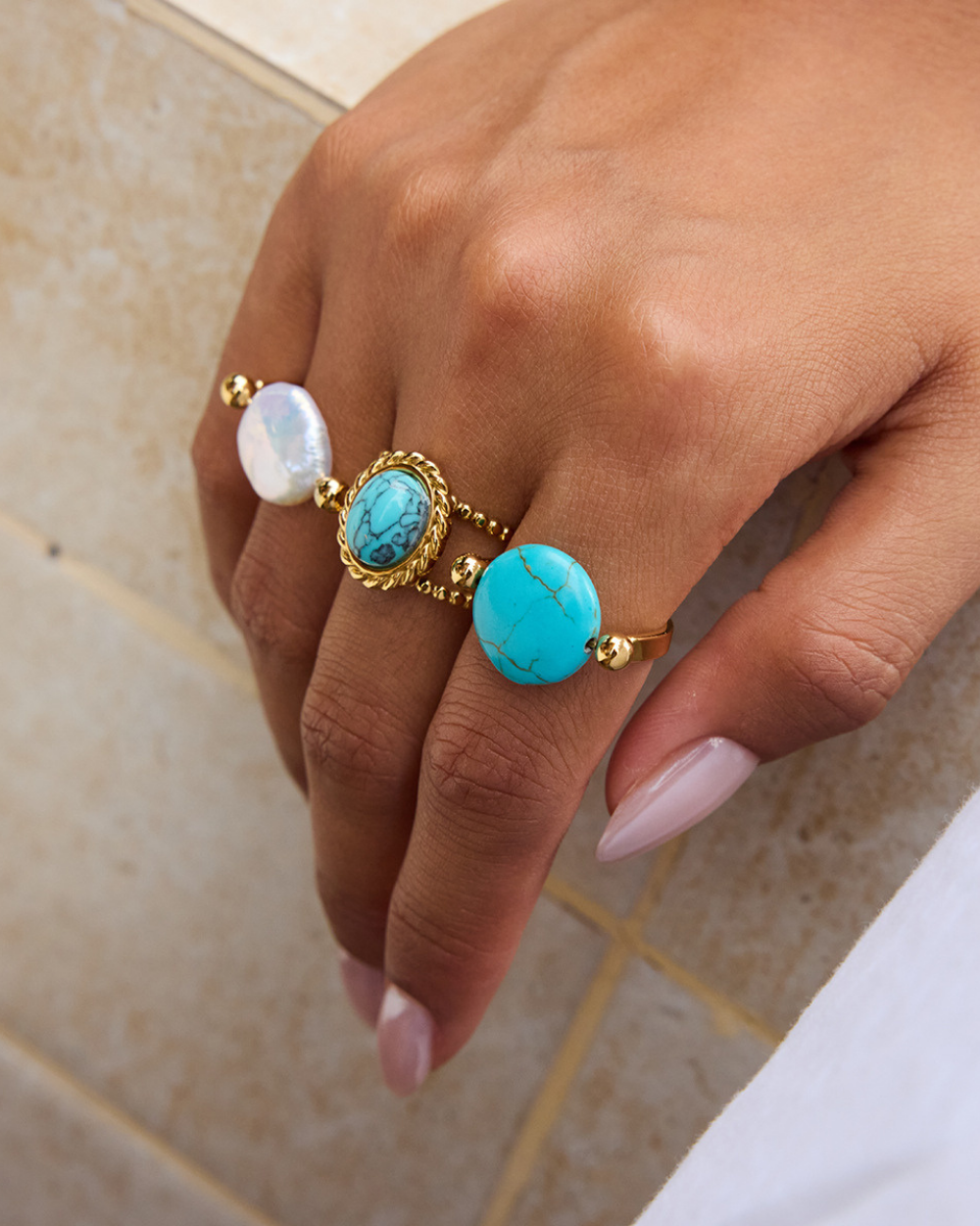 Hand wearing multiple rings with turquoise stones and pearls on a neutral background
