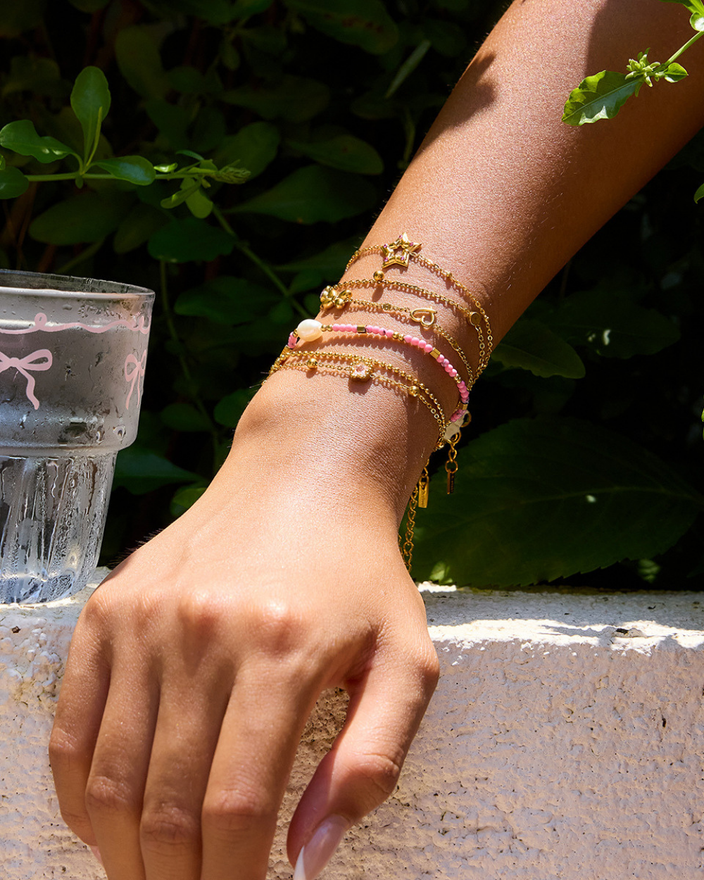 Hand wearing multiple gold and pink bracelets with a glass of water and greenery in the background