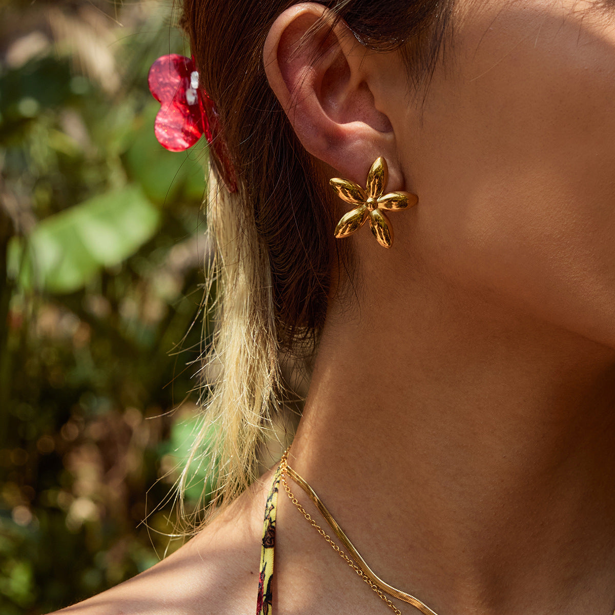 Gold five-petal earrings worn under sunlight near greenery