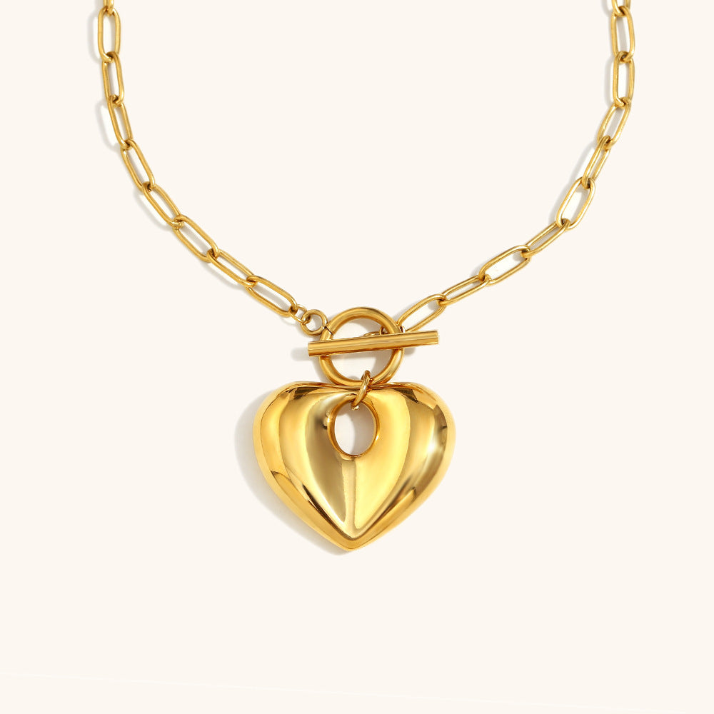 Close-up of a chunky gold heart pendant necklace with toggle clasp, shown on cream background