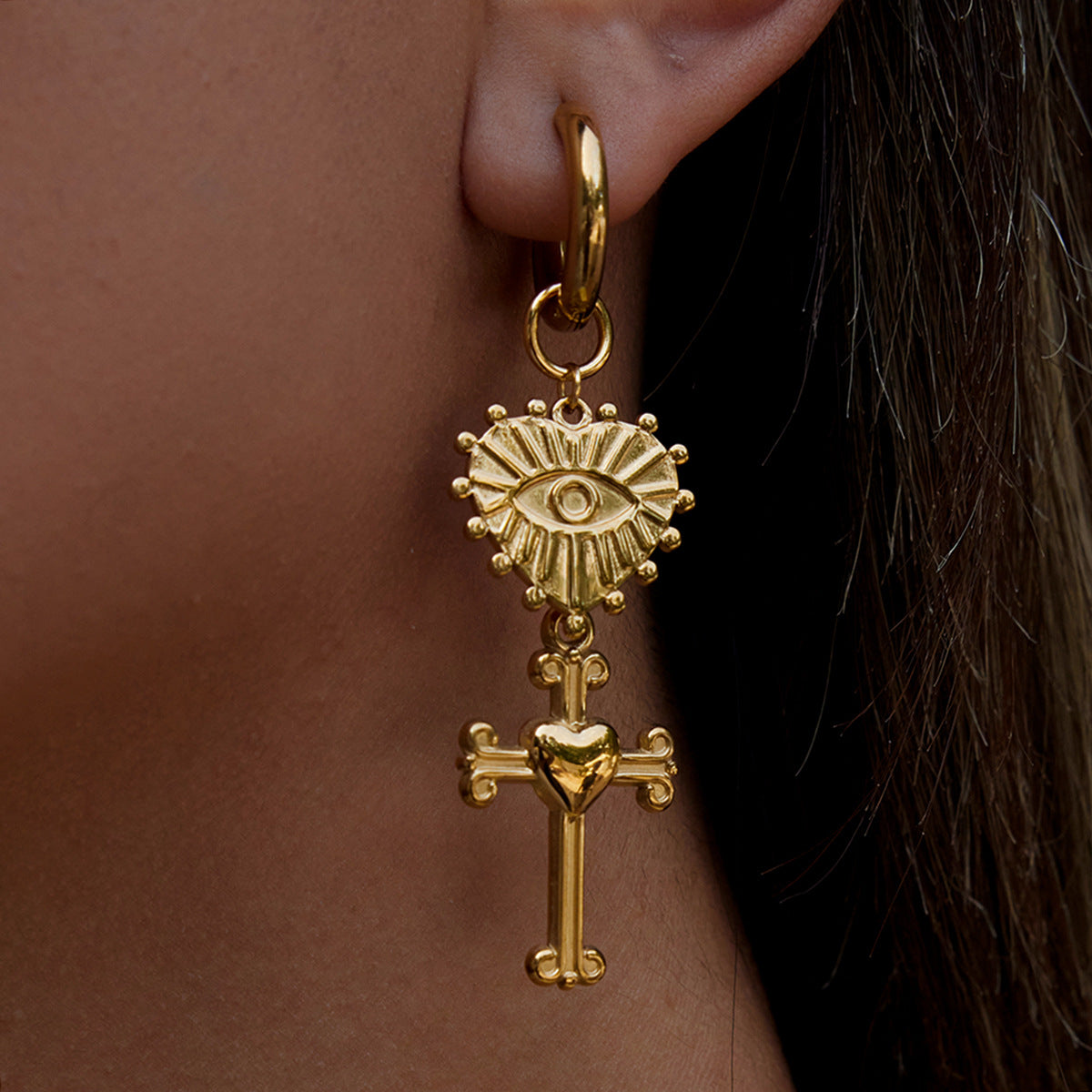 Gold earring with cross and heart design worn by a person.