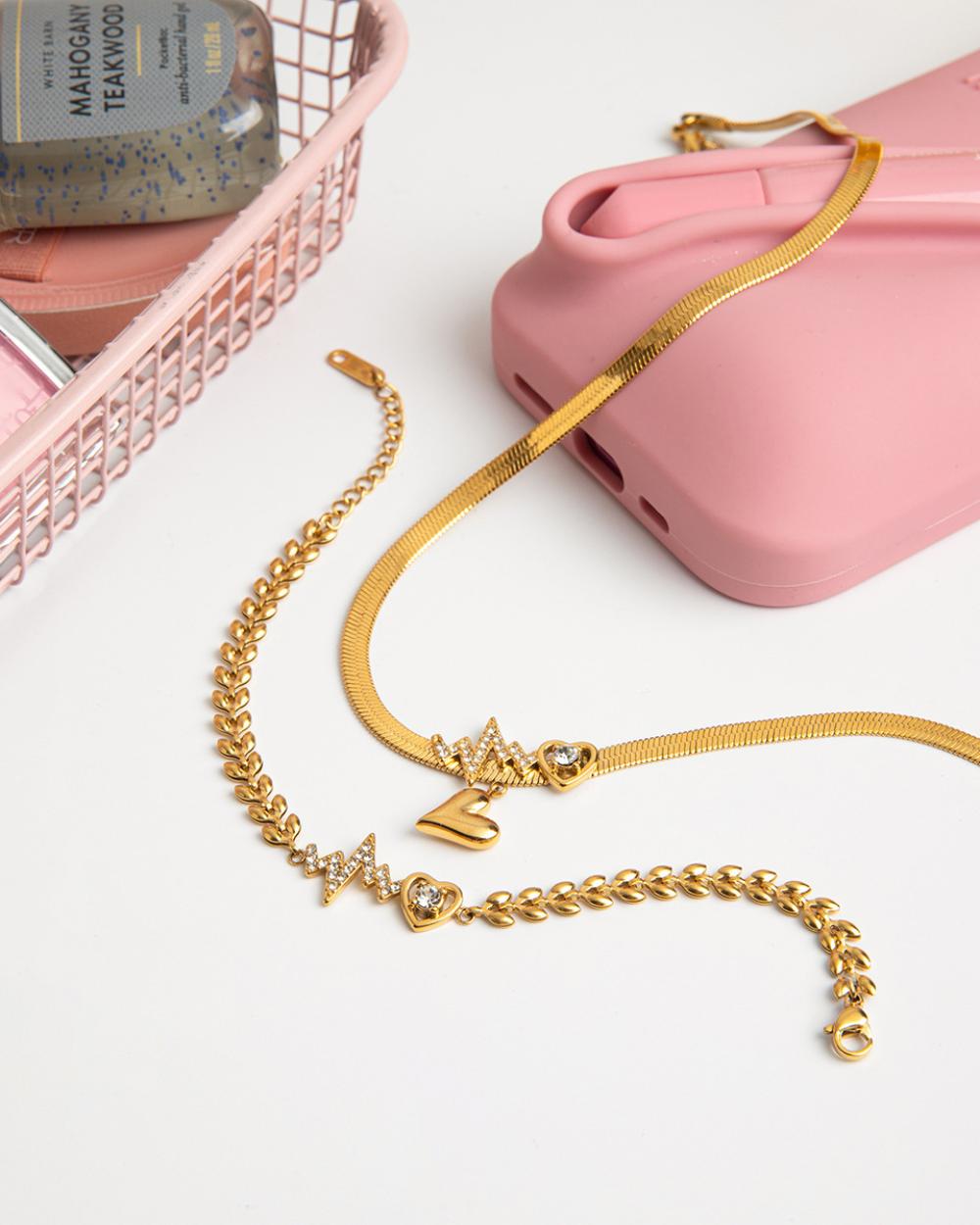 Gold necklace with star and heart charms on a white surface with pink phone case and basket in the background.