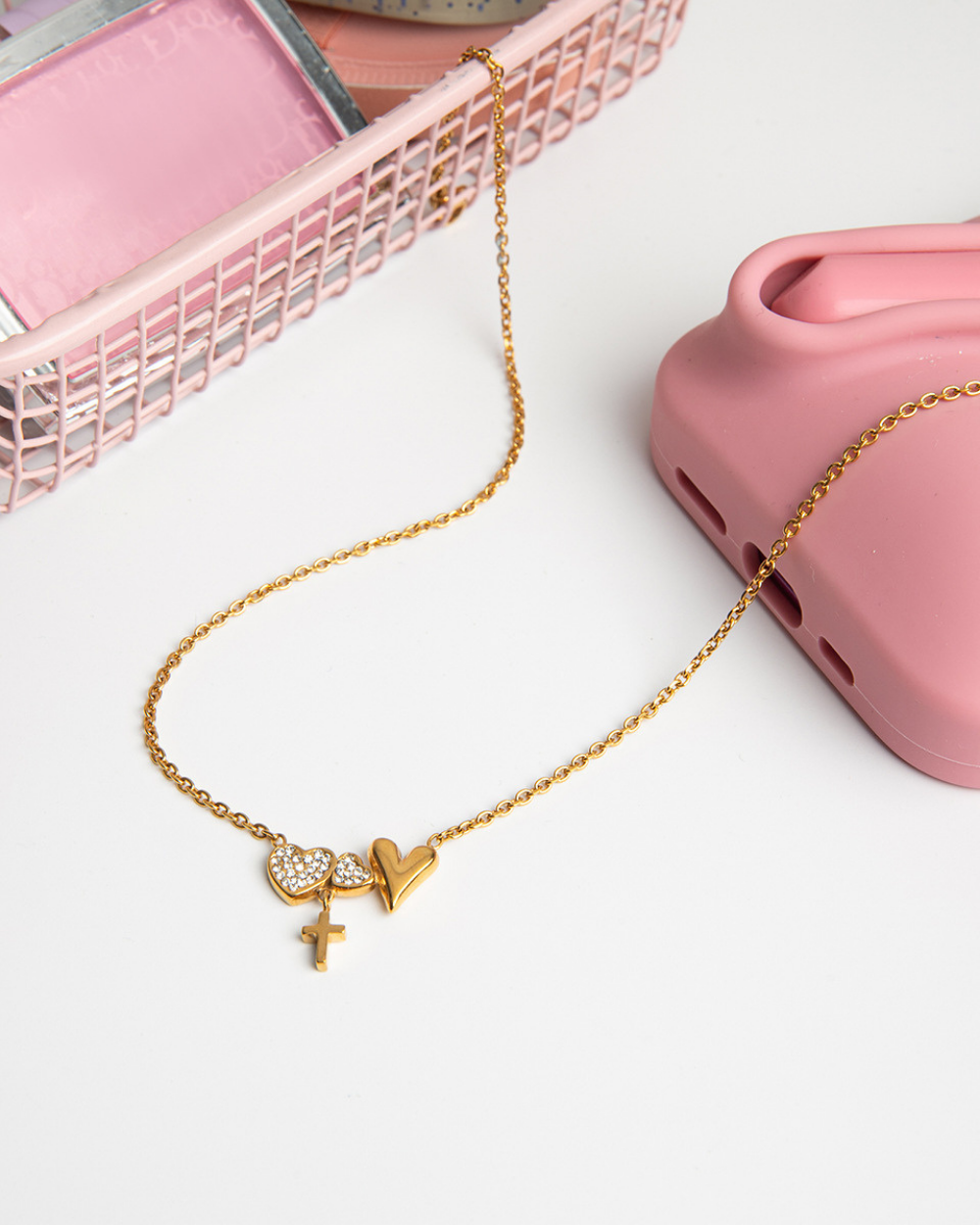 Gold necklace with heart, cross, and letter 'V' charms on a white surface with pink jewelry box and pink phone case.