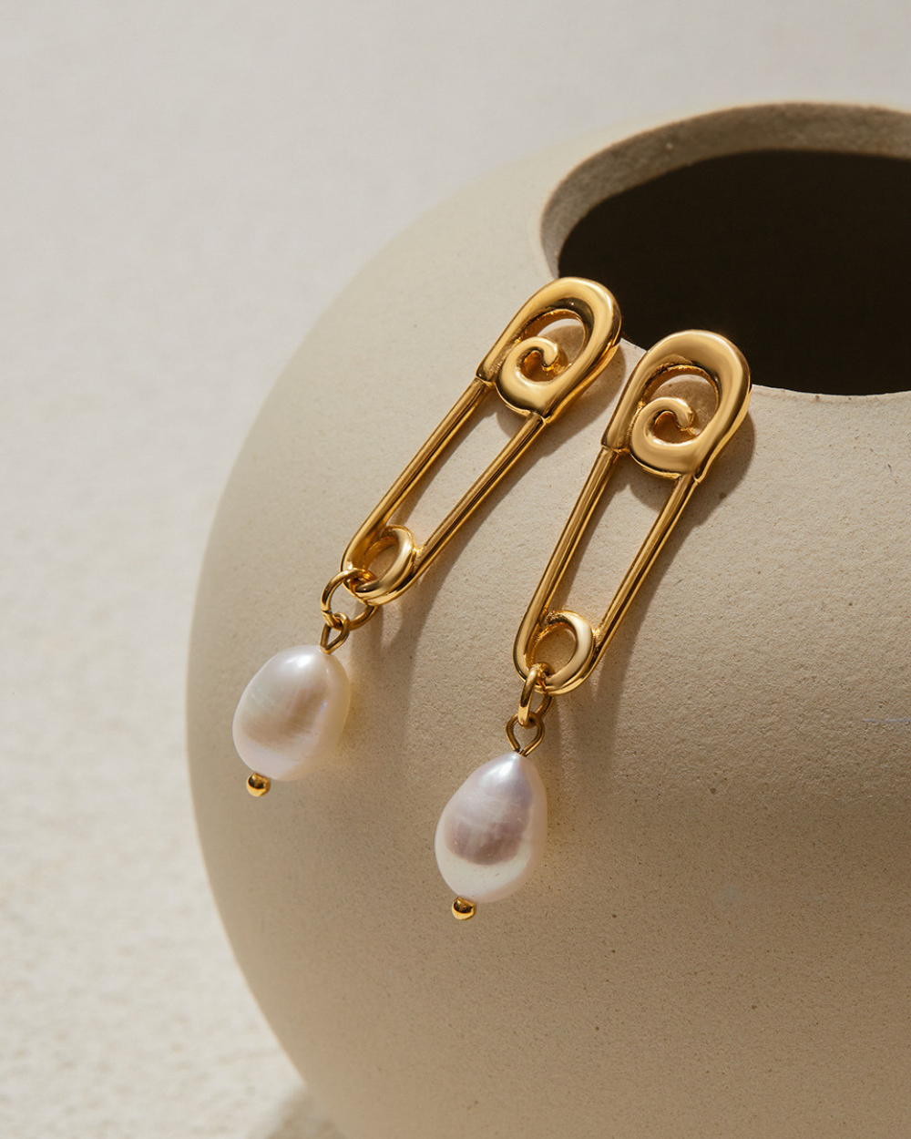 Gold safety pin earrings with pearl drops on a beige stand.