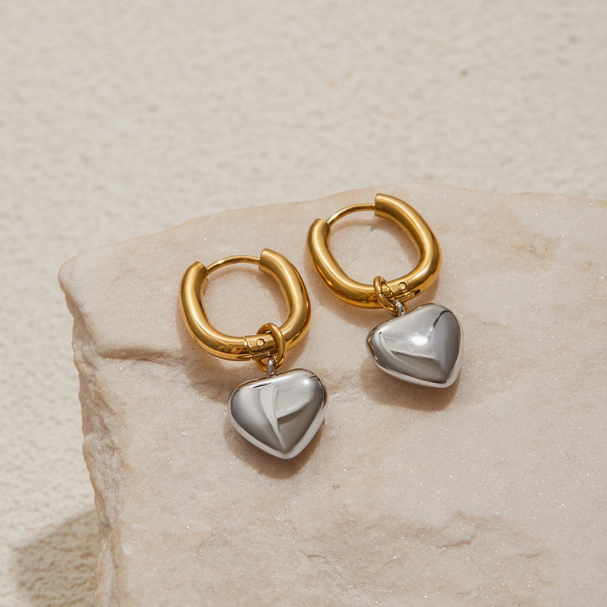 Gold hoop earrings with heart-shaped charms on a beige stone surface