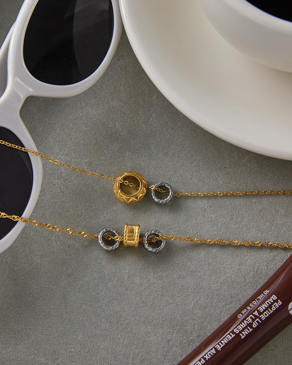 Two gold necklaces with intricate designs on a gray surface next to sunglasses and a cup.