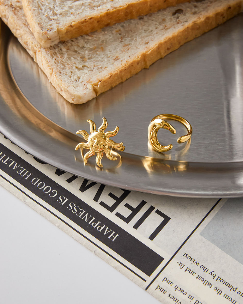 Gold sun and moon earrings on a metallic tray with slices of bread and a newspaper underneath.