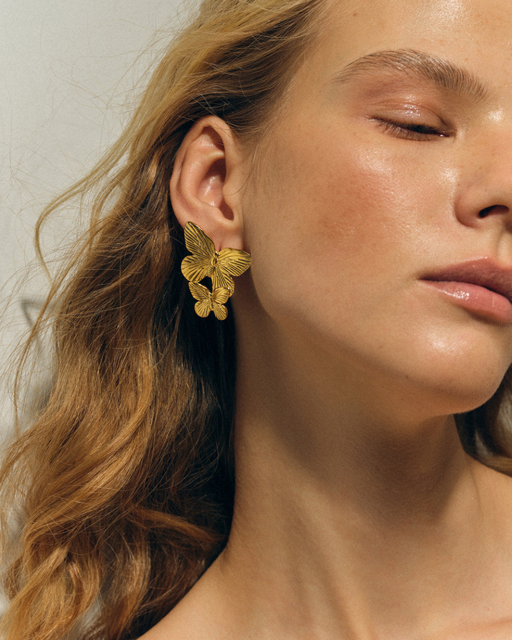 Close-up of a person wearing gold butterfly earrings with a neutral background
