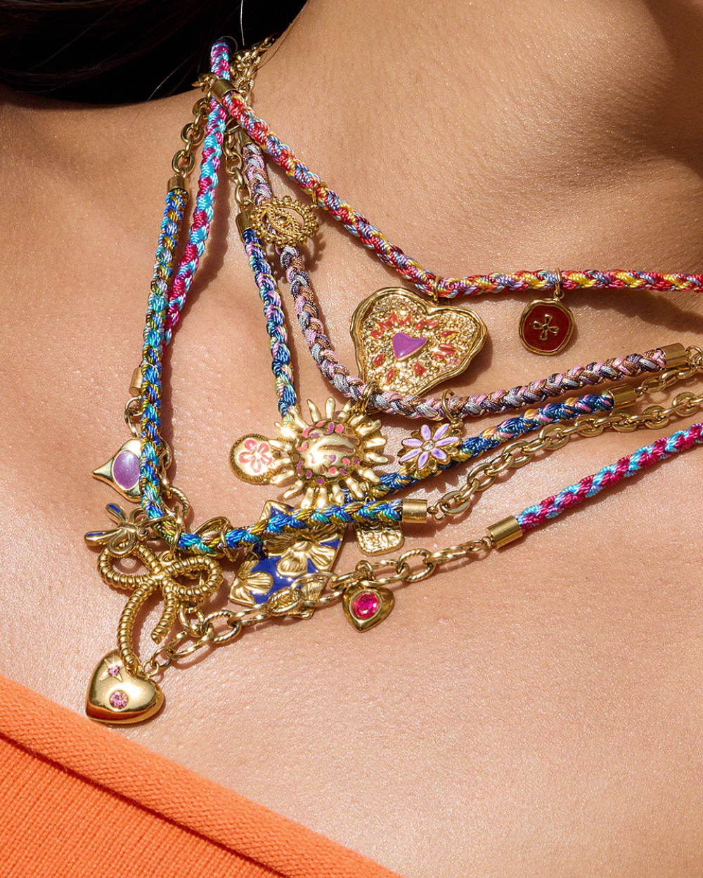 Colorful braided necklaces with gold hearts and charms on a person's neck against an orange fabric background.