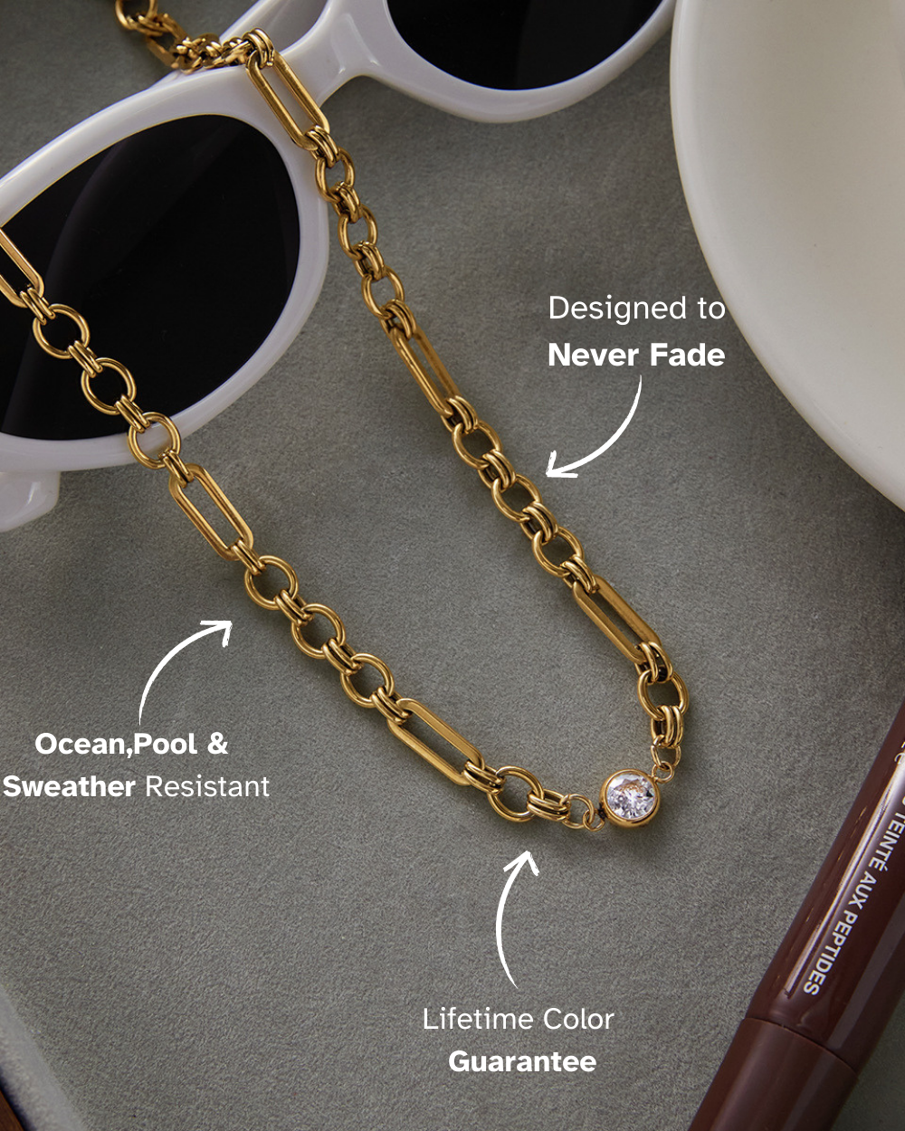 Gold chain with a gemstone, sunglasses, and a cup on a gray surface