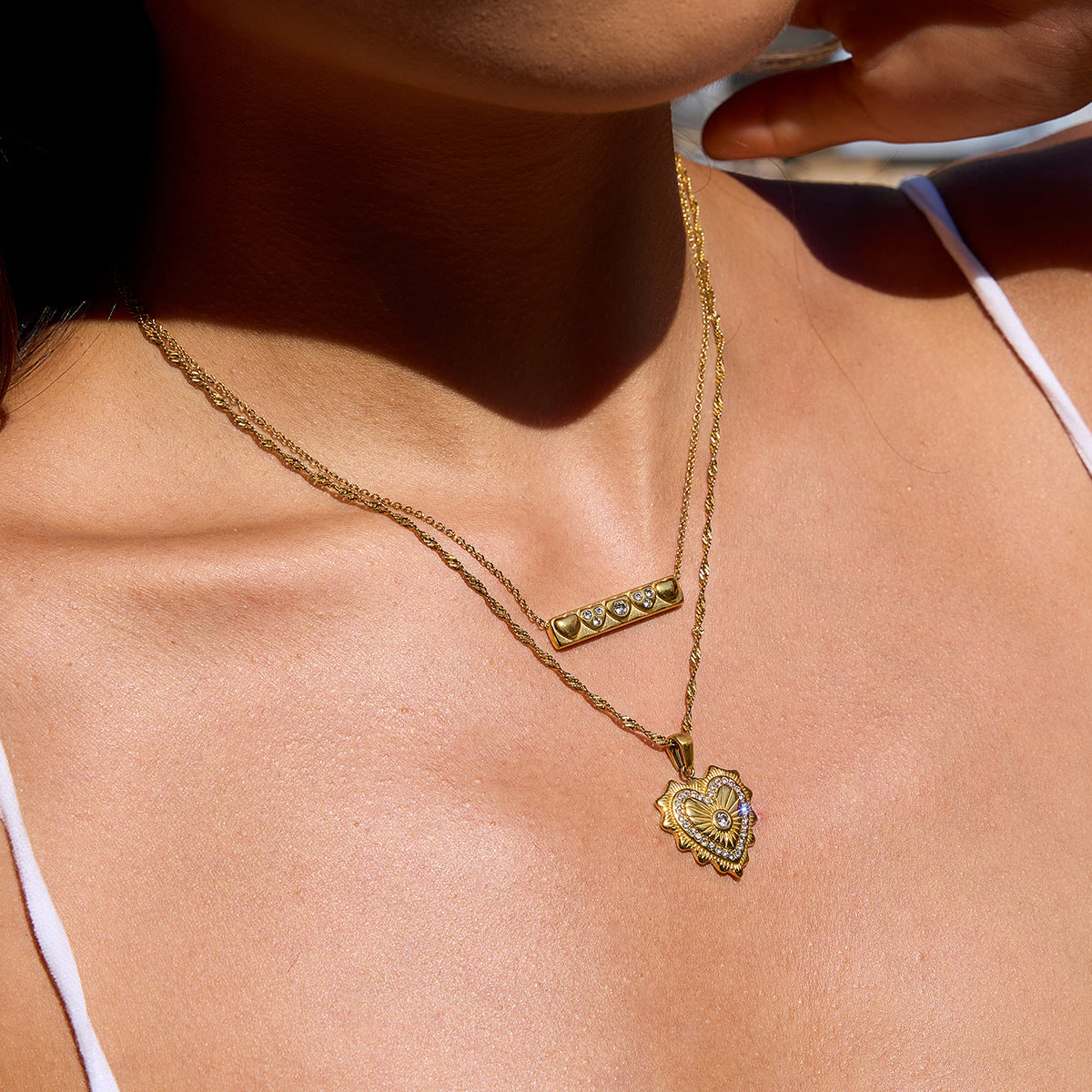 Model wearing layered gold necklaces with heart and bar pendants on sunlit skin