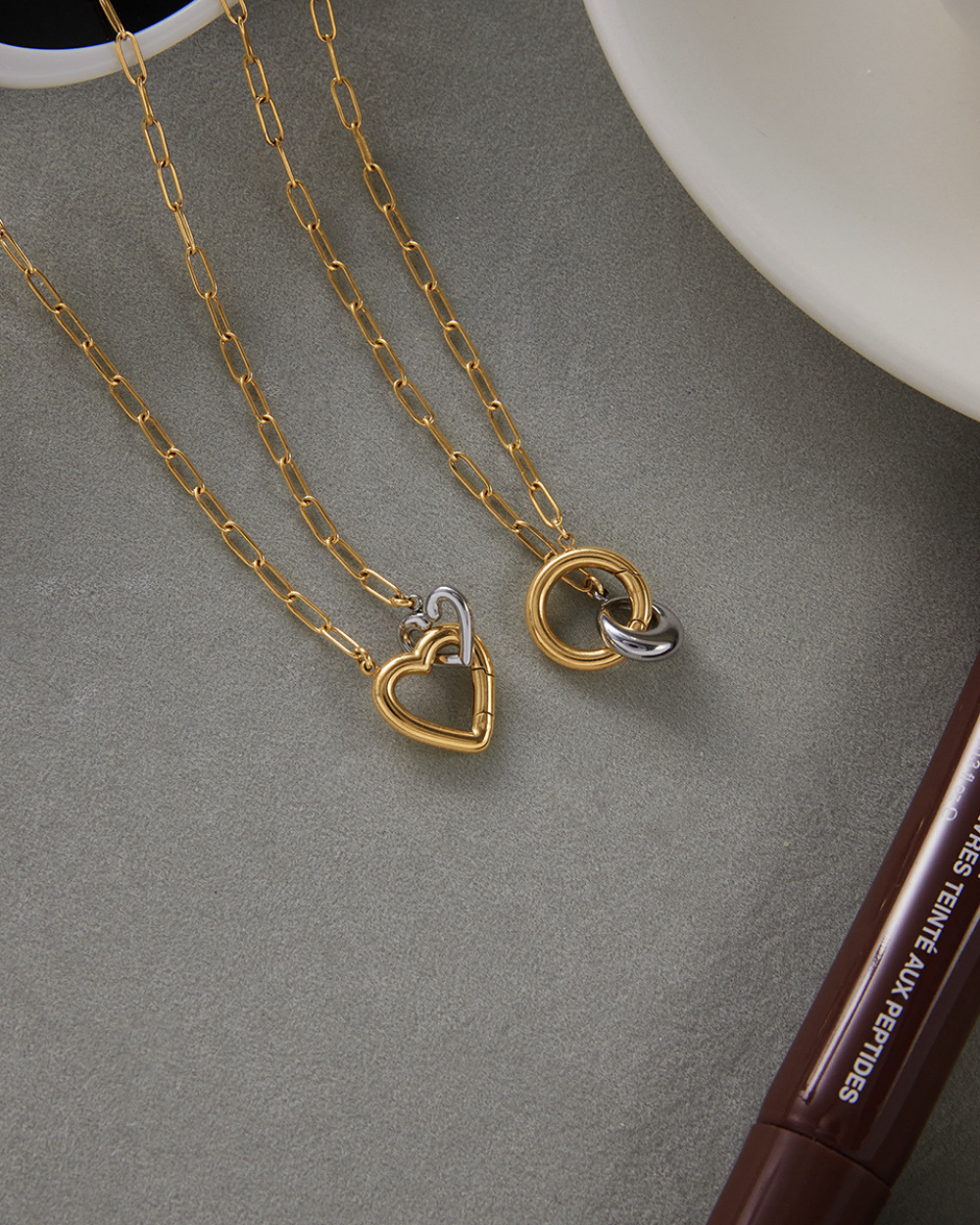 Two gold necklaces with heart and circular pendants on a gray surface.